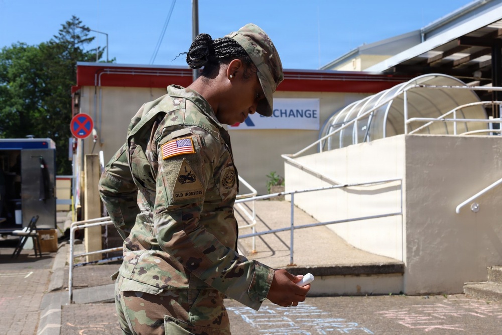 A U.S. Army soldier in OCP (Operational Camouflage Pattern) uniform is standing outdoors, looking down at a piece of chalk in their hand. The soldier's uniform displays a U.S. flag patch and an 'Old Ironsides' (1st Armored Division) unit patch. The setting appears to be a military installation, with an Army &amp; Air Force Exchange Service (AAFES) building visible in the background.