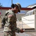 A U.S. Army soldier in OCP (Operational Camouflage Pattern) uniform is standing outdoors, looking down at a piece of chalk in their hand. The soldier's uniform displays a U.S. flag patch and an 'Old Ironsides' (1st Armored Division) unit patch. The setting appears to be a military installation, with an Army &amp; Air Force Exchange Service (AAFES) building visible in the background.