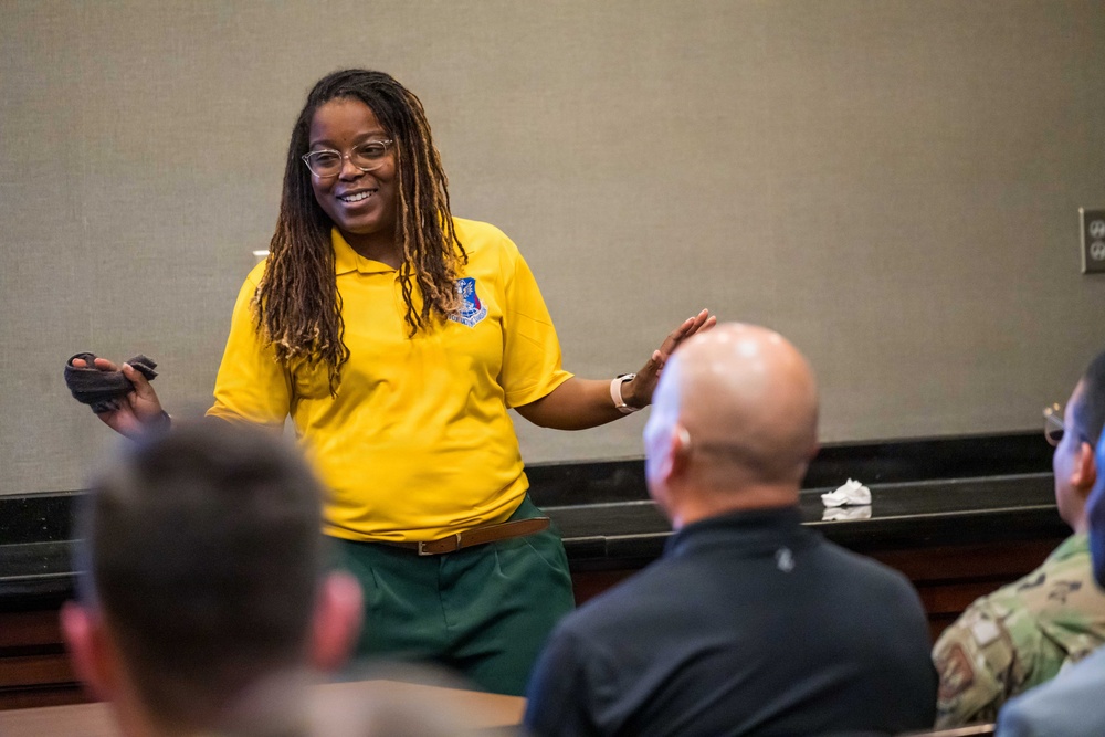 A woman in a yellow polo shirt with an emblem is presenting or speaking to a seated audience in what appears to be a conference or briefing room. She is smiling and gesturing with both hands, holding a small device in one hand, suggesting an engaging presentation. At least three audience members are visible from behind, including one in military camouflage uniform.