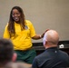 A woman in a yellow polo shirt with an emblem is presenting or speaking to a seated audience in what appears to be a conference or briefing room. She is smiling and gesturing with both hands, holding a small device in one hand, suggesting an engaging presentation. At least three audience members are visible from behind, including one in military camouflage uniform.