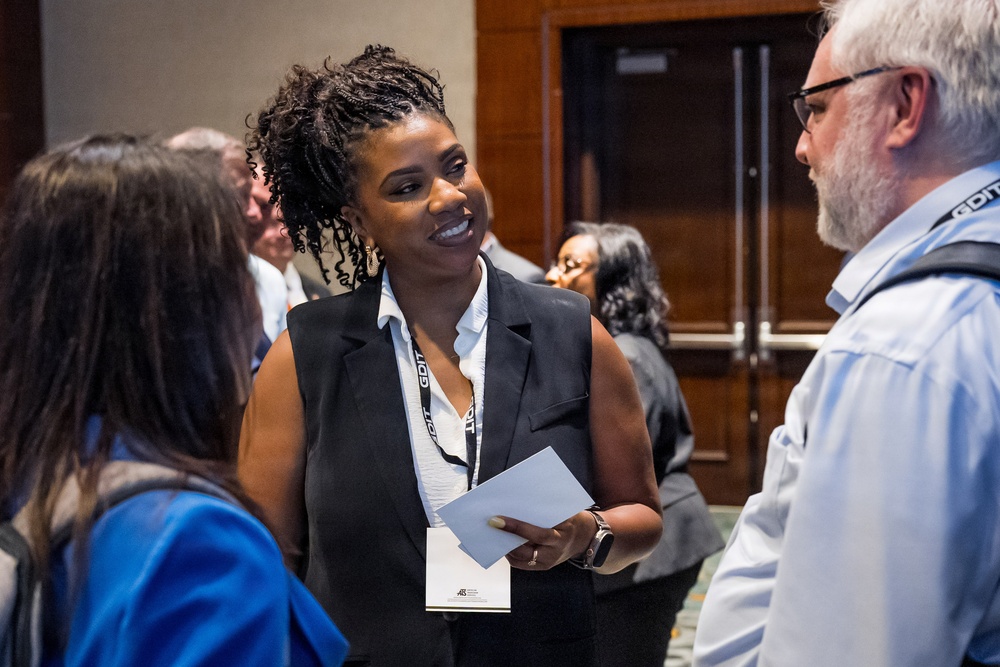 A professional networking event or conference where attendees are engaged in conversation. A woman in a black sleeveless blazer and white blouse, wearing a lanyard badge, is smiling and holding papers while speaking with others. The setting appears to be a hotel or convention center hallway or foyer.