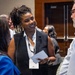 A professional networking event or conference where attendees are engaged in conversation. A woman in a black sleeveless blazer and white blouse, wearing a lanyard badge, is smiling and holding papers while speaking with others. The setting appears to be a hotel or convention center hallway or foyer.