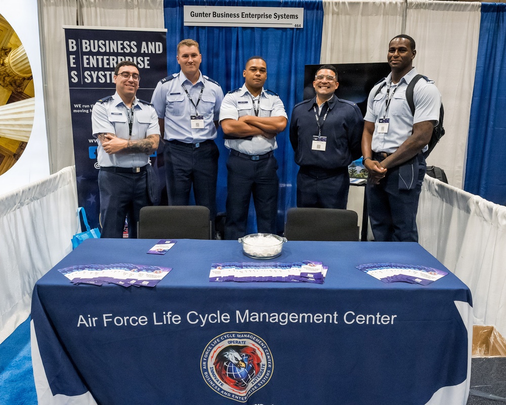 Five individuals, four in U.S. Air Force uniforms and one in civilian attire with a lanyard badge, pose at a booth for the Air Force Life Cycle Management Center (AFLCMC) at what appears to be a professional conference or trade show. The booth is associated with Gunter Business Enterprise Systems (BES), booth number 464. A navy blue tablecloth bearing the AFLCMC name and seal is displayed prominently in the foreground.