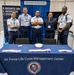 Five individuals, four in U.S. Air Force uniforms and one in civilian attire with a lanyard badge, pose at a booth for the Air Force Life Cycle Management Center (AFLCMC) at what appears to be a professional conference or trade show. The booth is associated with Gunter Business Enterprise Systems (BES), booth number 464. A navy blue tablecloth bearing the AFLCMC name and seal is displayed prominently in the foreground.
