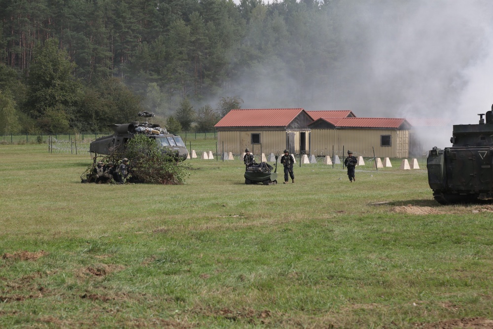 A military training exercise is underway on an open grassy field, featuring a downed or landed helicopter camouflaged with foliage, armed personnel moving across the field, and smoke rising near modular buildings in the background. An armored vehicle is visible on the right edge of the frame. The scene depicts a simulated combat or rescue scenario at what appears to be a military training facility.
