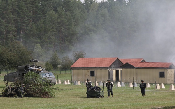 A military training exercise is underway on an open grassy field, featuring a downed or landed helicopter camouflaged with foliage, armed personnel moving across the field, and smoke rising near modular buildings in the background. An armored vehicle is visible on the right edge of the frame. The scene depicts a simulated combat or rescue scenario at what appears to be a military training facility.