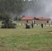A military training exercise is underway on an open grassy field, featuring a downed or landed helicopter camouflaged with foliage, armed personnel moving across the field, and smoke rising near modular buildings in the background. An armored vehicle is visible on the right edge of the frame. The scene depicts a simulated combat or rescue scenario at what appears to be a military training facility.