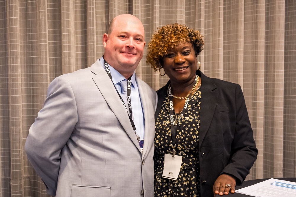 Two individuals are posing together for a photo at what appears to be a professional conference or corporate event. Both are wearing lanyards with 'GDIT' branding, suggesting affiliation with General Dynamics Information Technology. They are smiling and standing in front of a gray curtain backdrop.