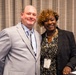 Two individuals are posing together for a photo at what appears to be a professional conference or corporate event. Both are wearing lanyards with 'GDIT' branding, suggesting affiliation with General Dynamics Information Technology. They are smiling and standing in front of a gray curtain backdrop.