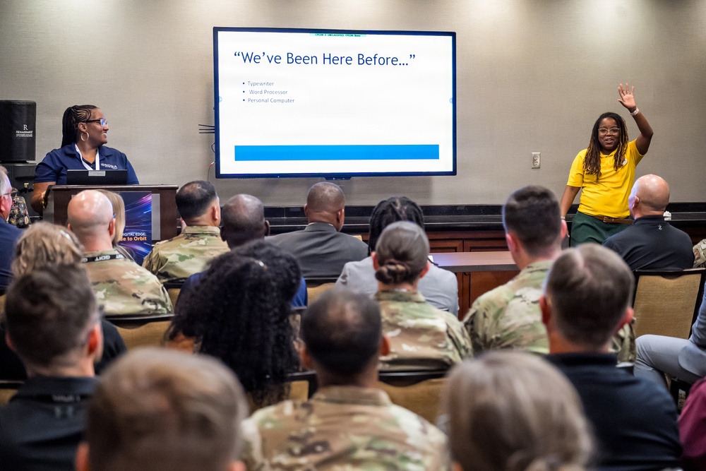 A presentation or briefing is taking place in a conference room setting, with a large display screen showing a slide titled 'We've Been Here Before...' listing Typewriter, Word Processor, and Personal Computer. The audience consists predominantly of military personnel in camouflage uniforms, along with some civilians. Two women appear to be co-presenting — one at a podium on the left in a blue shirt, and one in a yellow shirt on the right with her hand raised.