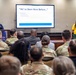 A presentation or briefing is taking place in a conference room setting, with a large display screen showing a slide titled 'We've Been Here Before...' listing Typewriter, Word Processor, and Personal Computer. The audience consists predominantly of military personnel in camouflage uniforms, along with some civilians. Two women appear to be co-presenting — one at a podium on the left in a blue shirt, and one in a yellow shirt on the right with her hand raised.
