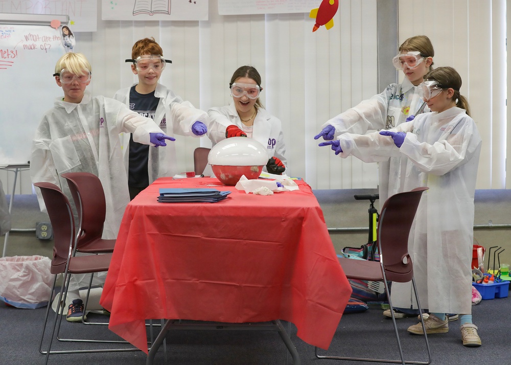 A group of five children are participating in a science demonstration or experiment in what appears to be a classroom setting. They are all wearing white lab coats, safety goggles, and purple gloves, gathered around a red-covered table with a large balloon or dome-shaped object on it. The children appear excited and engaged, with four of them pointing toward the central object while the middle child reacts with enthusiasm.