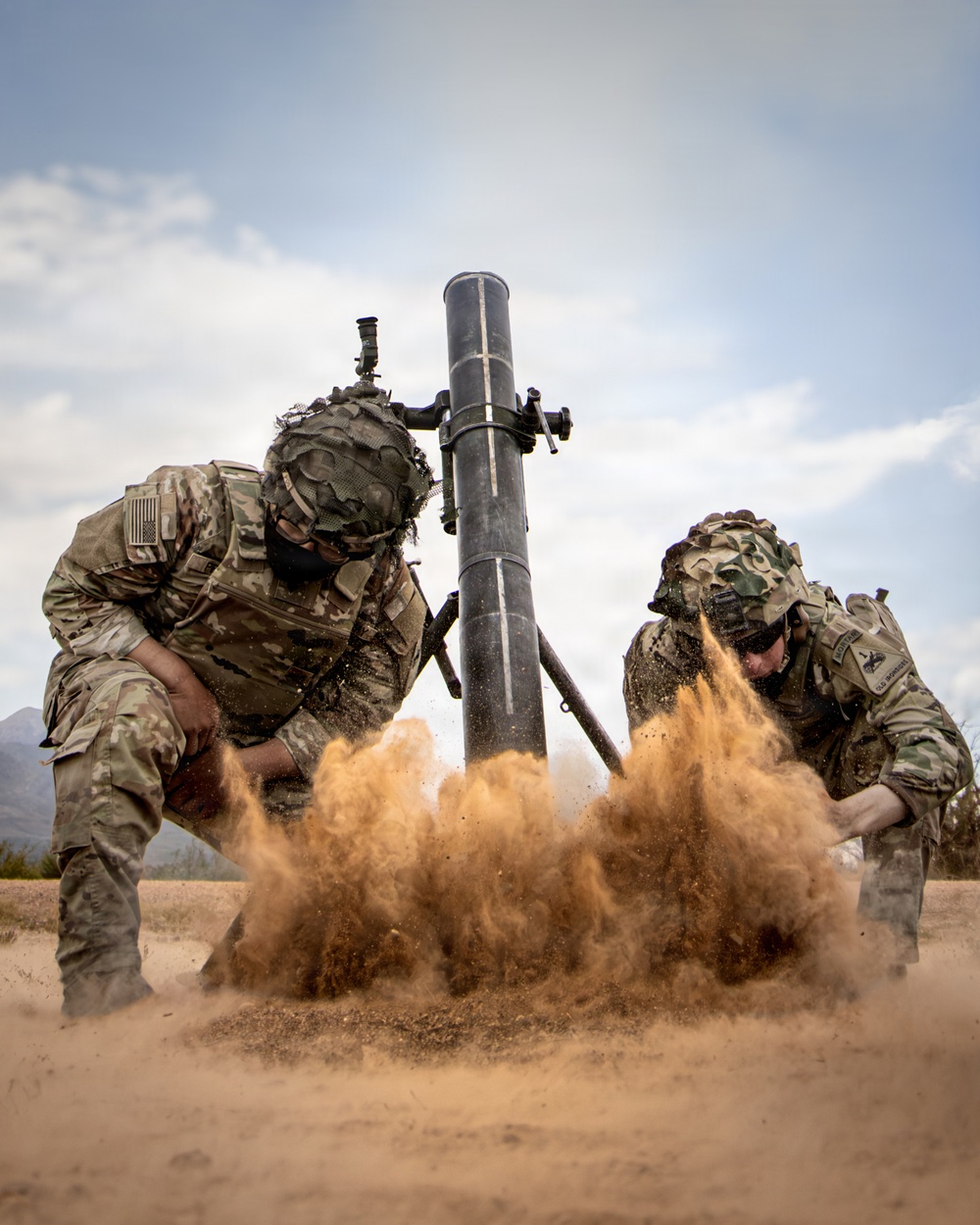 Two U.S. Army soldiers in full combat gear are firing a mortar system, captured at the moment of discharge with a dramatic explosion of sand and dust at the base plate. Both soldiers are wearing MultiCam camouflage uniforms with helmet covers and tactical vests, and are bracing against the blast effect. The image appears to be taken during a live-fire training exercise in an arid, desert-like environment.