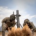 Two U.S. Army soldiers in full combat gear are firing a mortar system, captured at the moment of discharge with a dramatic explosion of sand and dust at the base plate. Both soldiers are wearing MultiCam camouflage uniforms with helmet covers and tactical vests, and are bracing against the blast effect. The image appears to be taken during a live-fire training exercise in an arid, desert-like environment.