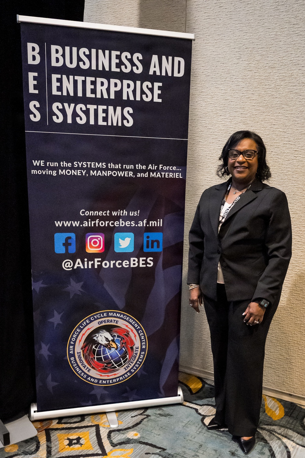 A woman in professional business attire stands beside a pull-up banner for the Air Force Business and Enterprise Systems (BES) directorate at what appears to be a conference or event. The banner displays the BES branding, website, social media handles, and the Air Force Life Cycle Management Center seal. The setting appears to be a hotel or convention center ballroom.