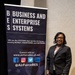 A woman in professional business attire stands beside a pull-up banner for the Air Force Business and Enterprise Systems (BES) directorate at what appears to be a conference or event. The banner displays the BES branding, website, social media handles, and the Air Force Life Cycle Management Center seal. The setting appears to be a hotel or convention center ballroom.