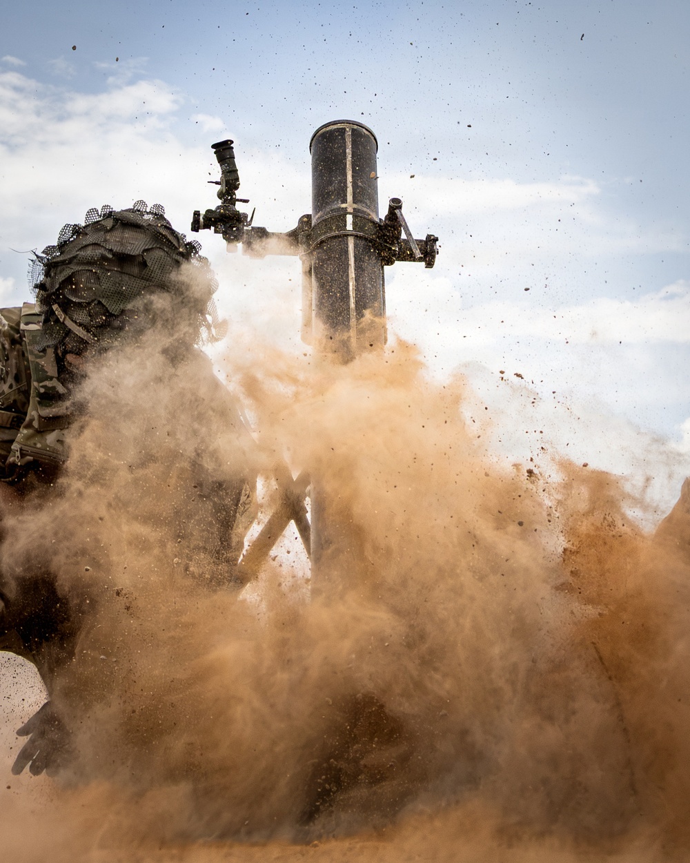 A mortar system is captured at the moment of firing, with a large cloud of displaced sand and dust erupting from the base plate impact. A soldier in camouflage gear with a helmet cover is visible on the left side of the frame. The image was taken at ground level in an arid desert environment, capturing the dramatic blast effect.