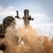 A mortar system is captured at the moment of firing, with a large cloud of displaced sand and dust erupting from the base plate impact. A soldier in camouflage gear with a helmet cover is visible on the left side of the frame. The image was taken at ground level in an arid desert environment, capturing the dramatic blast effect.