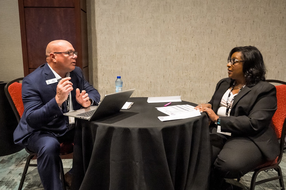 Two professionally dressed individuals are seated across from each other at a round table covered with a black tablecloth, engaged in what appears to be a formal meeting or interview. The man on the left is speaking and gesturing with his hands while the woman on the right listens attentively with documents in front of her. The setting appears to be a hotel conference or event room.