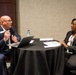Two professionally dressed individuals are seated across from each other at a round table covered with a black tablecloth, engaged in what appears to be a formal meeting or interview. The man on the left is speaking and gesturing with his hands while the woman on the right listens attentively with documents in front of her. The setting appears to be a hotel conference or event room.