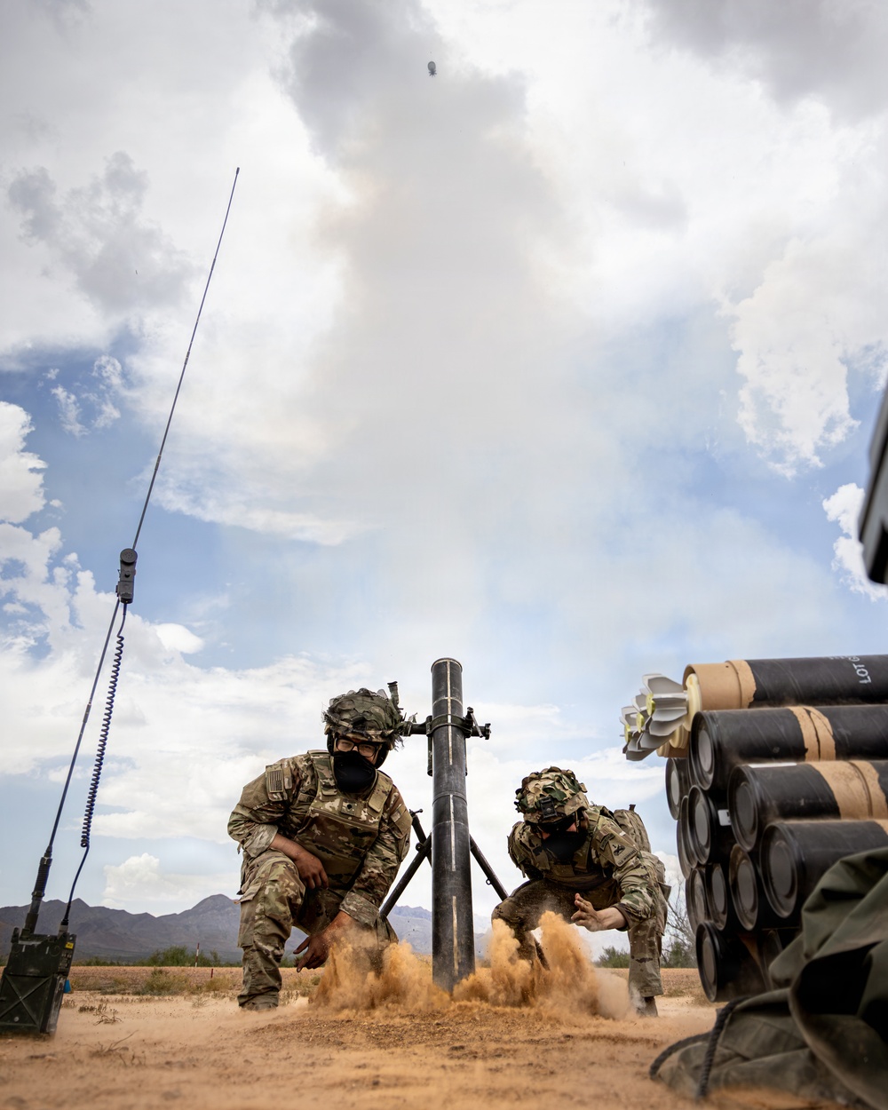 Two soldiers in full combat gear are operating a mortar system in an arid desert training environment. A mortar round is visibly in flight above the tube, with sand and dust being displaced by the blast. Multiple mortar rounds are staged on the right side, and a radio antenna is visible to the left.