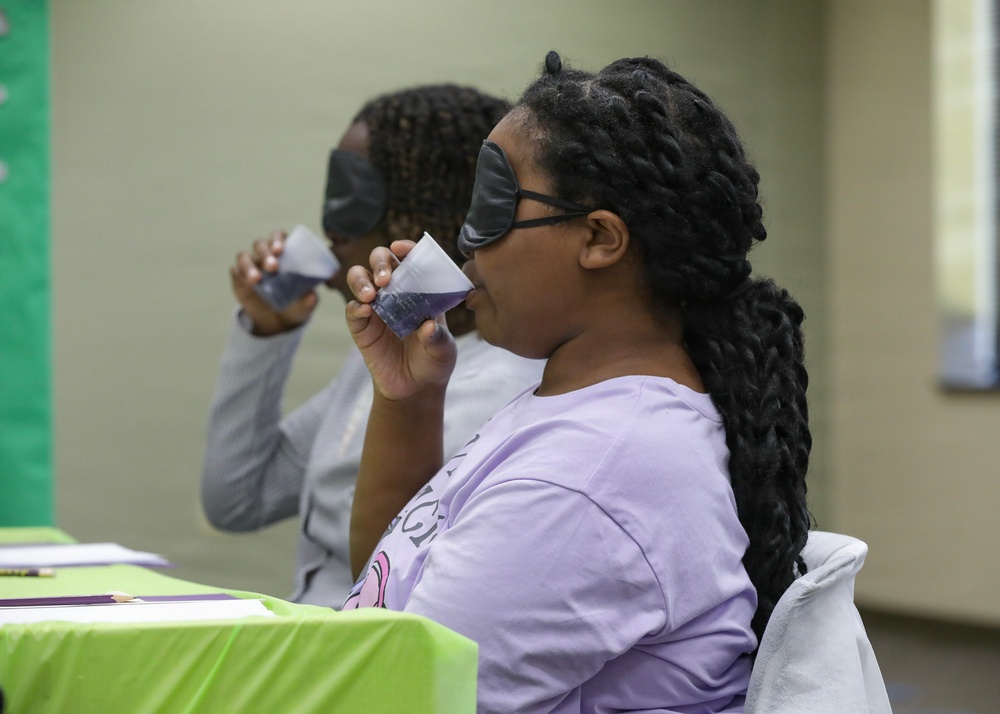 Two individuals are seated at a table covered with a bright green tablecloth, participating in what appears to be a blind taste test activity. The foreground participant is wearing a black sleep mask over her eyes and a lavender t-shirt, drinking from a small plastic cup. A second participant in the background is similarly drinking from a cup, suggesting a structured tasting event or competition.