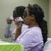 Two individuals are seated at a table covered with a bright green tablecloth, participating in what appears to be a blind taste test activity. The foreground participant is wearing a black sleep mask over her eyes and a lavender t-shirt, drinking from a small plastic cup. A second participant in the background is similarly drinking from a cup, suggesting a structured tasting event or competition.