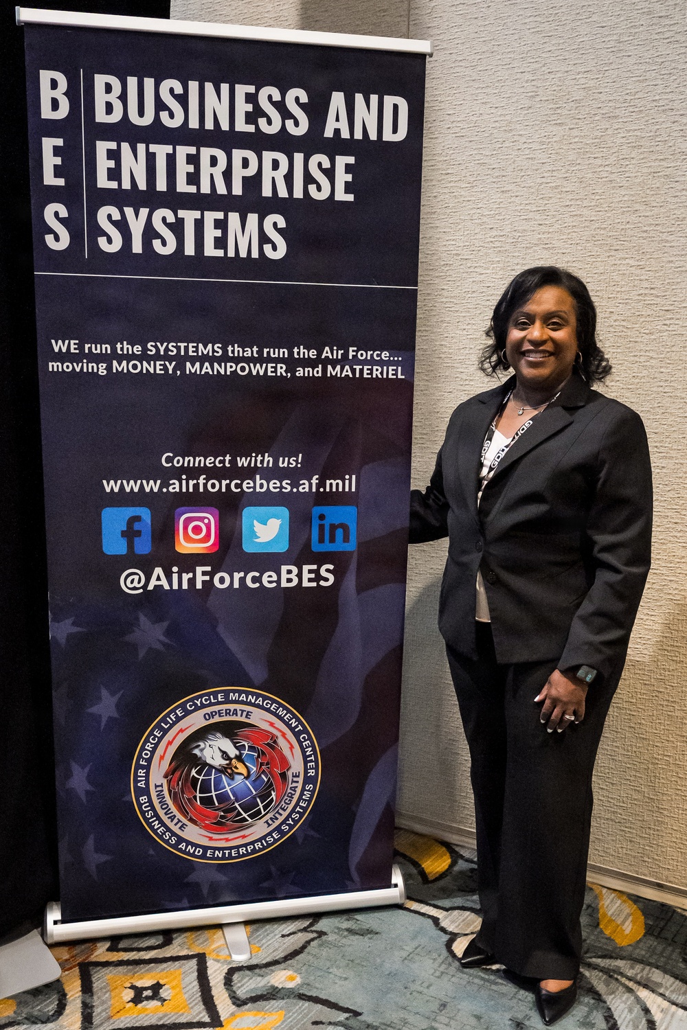 A woman in professional business attire stands beside a pull-up banner for 'Business and Enterprise Systems' (BES), an Air Force Life Cycle Management Center organization. She is smiling and posing for the photo in what appears to be a conference or event venue. The banner displays social media handles, a website URL, and the organization's seal.