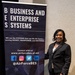 A woman in professional business attire stands beside a pull-up banner for 'Business and Enterprise Systems' (BES), an Air Force Life Cycle Management Center organization. She is smiling and posing for the photo in what appears to be a conference or event venue. The banner displays social media handles, a website URL, and the organization's seal.