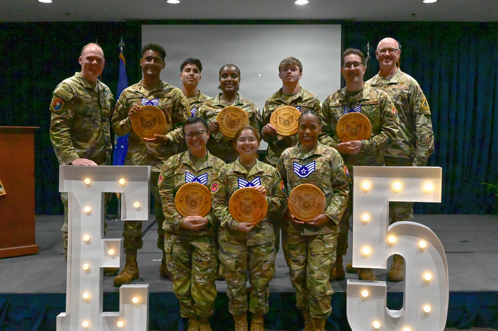 A group of U.S. Air Force military personnel pose for a formal group photo at what appears to be a promotion ceremony. Several members are holding circular wooden plaques, likely commemorating their promotion to Staff Sergeant or Technical Sergeant (E-5). The group is posed in front of large illuminated letter/number decorations spelling 'E5'.