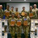 A group of U.S. Air Force military personnel pose for a formal group photo at what appears to be a promotion ceremony. Several members are holding circular wooden plaques, likely commemorating their promotion to Staff Sergeant or Technical Sergeant (E-5). The group is posed in front of large illuminated letter/number decorations spelling 'E5'.