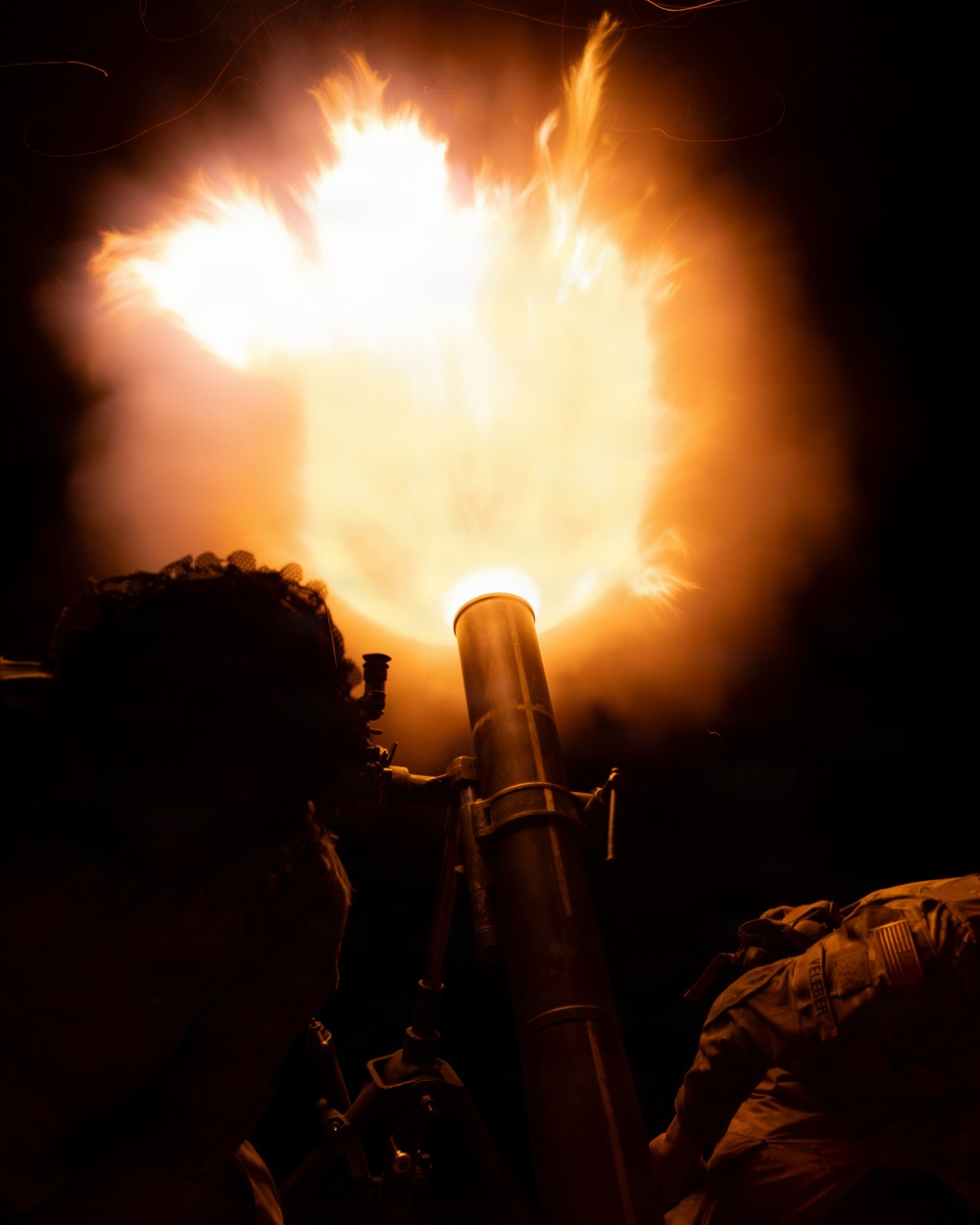 A nighttime military mortar firing captured at the moment of discharge, producing a large muzzle blast fireball illuminating the scene in orange and white light. Two soldiers in military uniforms are silhouetted against the intense muzzle flash. The image appears to be a training or operational exercise conducted in darkness.