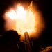 A nighttime military mortar firing captured at the moment of discharge, producing a large muzzle blast fireball illuminating the scene in orange and white light. Two soldiers in military uniforms are silhouetted against the intense muzzle flash. The image appears to be a training or operational exercise conducted in darkness.