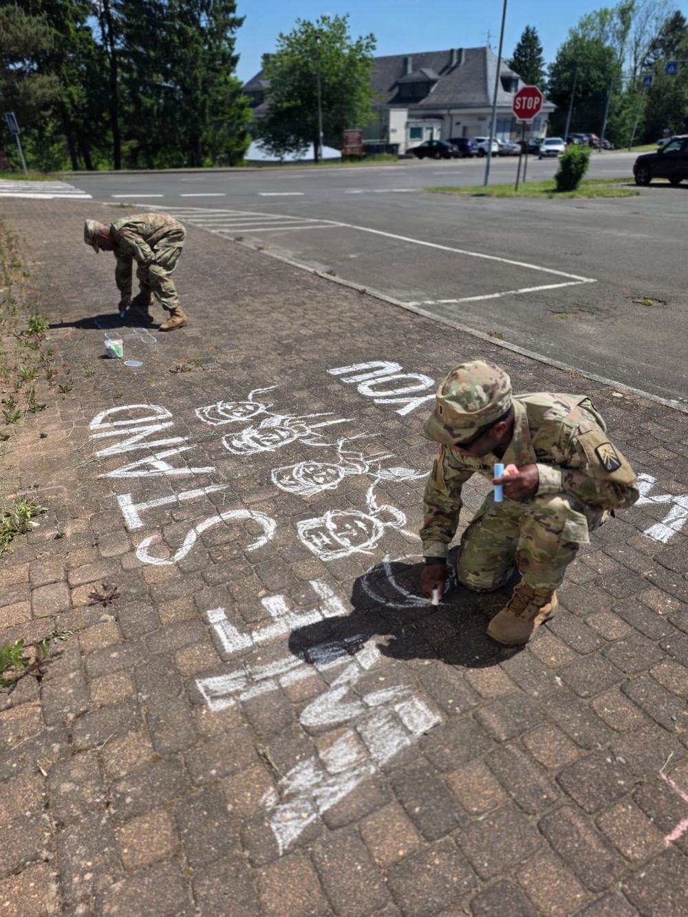 Two soldiers in U.S. Army camouflage (OCP) uniforms are crouching on a brick-paved sidewalk or plaza, writing and drawing with chalk. The chalk artwork includes text reading 'STAND' and what appears to be 'SEMI' or similar lettering, along with drawn figures resembling children or people. The setting appears to be a military installation or base, with a stop sign, buildings, parked vehicles, and trees visible in the background.