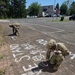 Two soldiers in U.S. Army camouflage (OCP) uniforms are crouching on a brick-paved sidewalk or plaza, writing and drawing with chalk. The chalk artwork includes text reading 'STAND' and what appears to be 'SEMI' or similar lettering, along with drawn figures resembling children or people. The setting appears to be a military installation or base, with a stop sign, buildings, parked vehicles, and trees visible in the background.