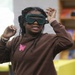 A young student is putting on green safety goggles, likely in preparation for a science experiment or lab activity. She is wearing a brown hoodie with a pink butterfly graphic and multiple bracelets. The setting appears to be a classroom or school library environment.