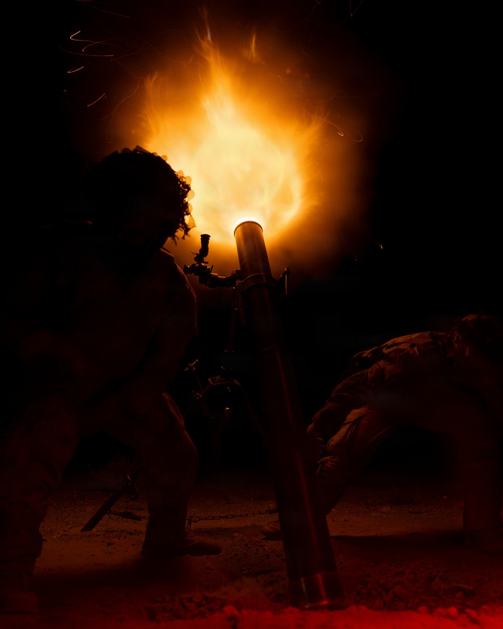 A nighttime image showing silhouetted figures alongside what appears to be a mortar or artillery piece being fired, producing a large muzzle blast of flame and light. The scene is dramatically lit solely by the intense orange and white fireball emanating from the barrel. The high-contrast lighting renders the human figures and equipment almost entirely in silhouette against the dark background.