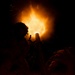A nighttime image showing silhouetted figures alongside what appears to be a mortar or artillery piece being fired, producing a large muzzle blast of flame and light. The scene is dramatically lit solely by the intense orange and white fireball emanating from the barrel. The high-contrast lighting renders the human figures and equipment almost entirely in silhouette against the dark background.