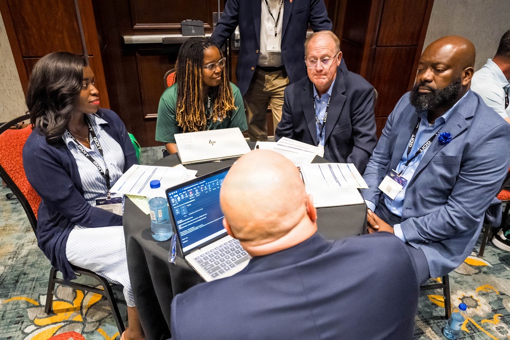 A group of approximately six professionals are gathered around a round table covered with a black tablecloth in what appears to be a hotel conference room or meeting space. They are engaged in a collaborative discussion or working session, with documents, papers, and at least one open laptop visible on the table. All visible participants are wearing conference lanyards/badges, suggesting this is part of a formal professional event.