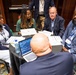 A group of approximately six professionals are gathered around a round table covered with a black tablecloth in what appears to be a hotel conference room or meeting space. They are engaged in a collaborative discussion or working session, with documents, papers, and at least one open laptop visible on the table. All visible participants are wearing conference lanyards/badges, suggesting this is part of a formal professional event.