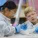 Two children are engaged in a science dissection activity, likely in a classroom setting. Both are wearing protective gear including lab coats, safety goggles, and latex gloves. The girl in the foreground is smiling and using forceps/pliers to examine a specimen, while the boy in the background observes.