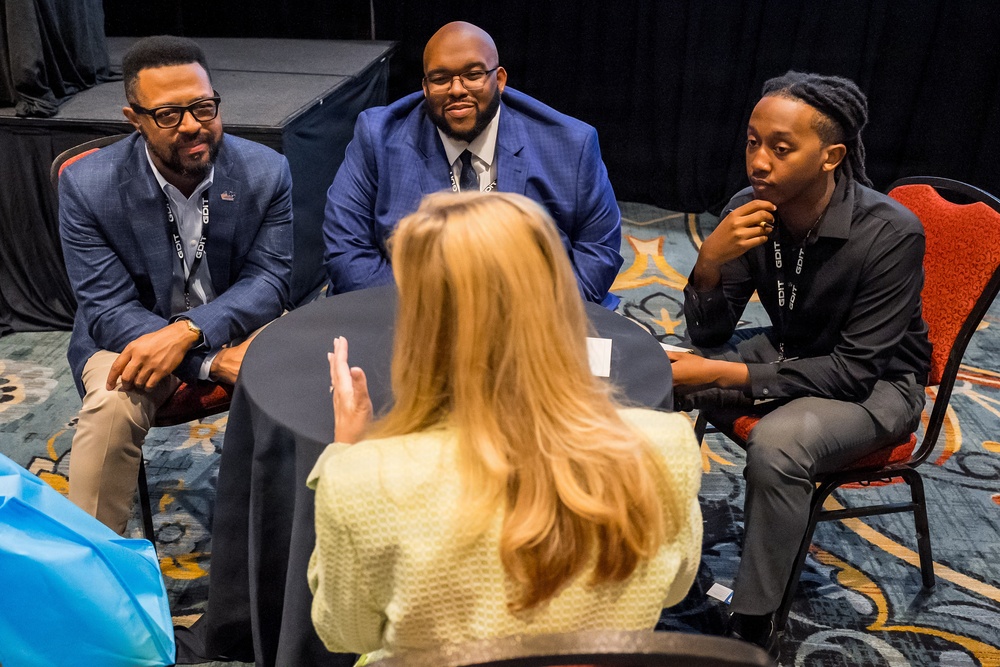 Four individuals are engaged in a small group discussion around a round table covered with a black tablecloth, in what appears to be a conference or professional event setting. Three participants are visible from the front — two men in blue blazers and one younger man in black — while a fourth person with blonde hair is seen from behind, gesturing with her hand as she speaks. Lanyards with 'GDIT' branding are visible on multiple attendees.