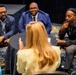 Four individuals are engaged in a small group discussion around a round table covered with a black tablecloth, in what appears to be a conference or professional event setting. Three participants are visible from the front — two men in blue blazers and one younger man in black — while a fourth person with blonde hair is seen from behind, gesturing with her hand as she speaks. Lanyards with 'GDIT' branding are visible on multiple attendees.