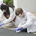 Two children are engaged in an outdoor science activity, wearing full protective gear including white lab coats, safety goggles, and purple nitrile gloves. They are crouched down on a paved surface, handling small white objects or containers as part of what appears to be a structured educational science experiment. A sign in the background references science and chemistry, suggesting this is an organized educational event.