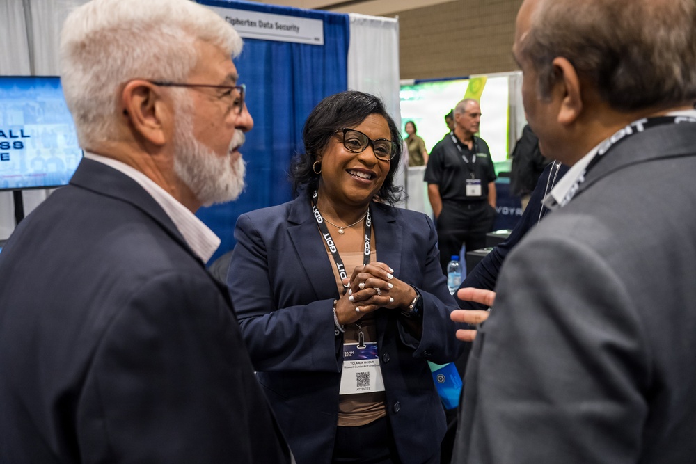 Three professionals are engaged in conversation at what appears to be a technology conference or trade show expo floor. The central figure is a woman wearing a navy blazer and conference badge, smiling warmly while speaking with two men. In the background, additional attendees and vendor booths, including one labeled 'Ciphertex Data Security,' are visible.