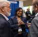 Three professionals are engaged in conversation at what appears to be a technology conference or trade show expo floor. The central figure is a woman wearing a navy blazer and conference badge, smiling warmly while speaking with two men. In the background, additional attendees and vendor booths, including one labeled 'Ciphertex Data Security,' are visible.