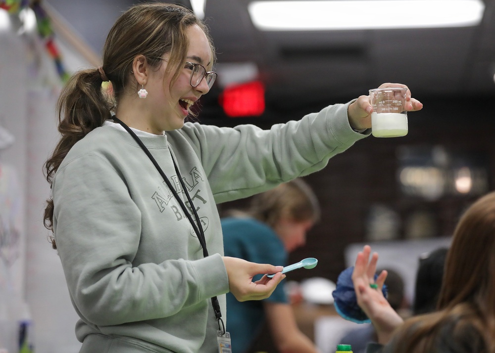 A young woman wearing a light gray sweatshirt and glasses is enthusiastically holding up a glass beaker containing a milky/greenish liquid during what appears to be a science activity or educational event. She is smiling broadly and holding a small blue spoon in her other hand, suggesting she is mixing or measuring substances. The setting appears to be an indoor educational or community event space with other participants visible in the blurred background.