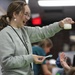 A young woman wearing a light gray sweatshirt and glasses is enthusiastically holding up a glass beaker containing a milky/greenish liquid during what appears to be a science activity or educational event. She is smiling broadly and holding a small blue spoon in her other hand, suggesting she is mixing or measuring substances. The setting appears to be an indoor educational or community event space with other participants visible in the blurred background.
