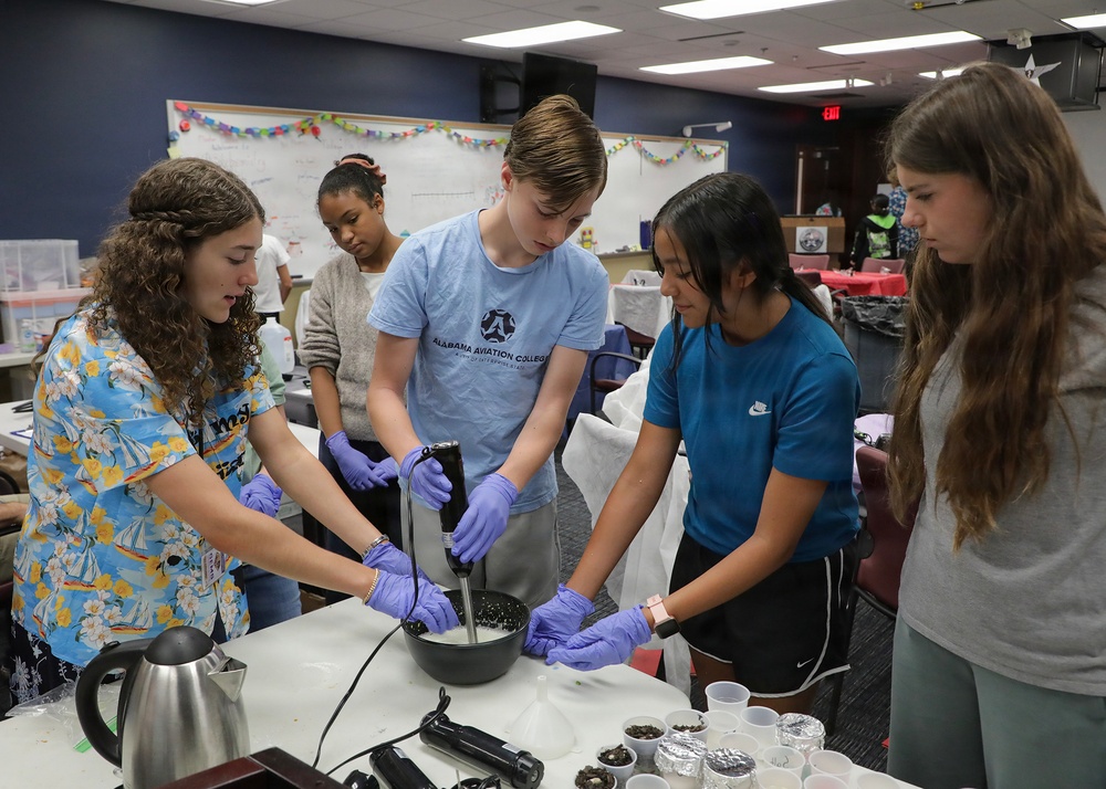 A group of young students are engaged in a hands-on science or culinary activity in what appears to be a classroom or educational facility. Three students wearing purple latex gloves are actively working with a hand blender in a bowl, while others observe. The setting includes a whiteboard, decorations, and various materials on the table suggesting an organized educational workshop.