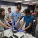 A group of young students are engaged in a hands-on science or culinary activity in what appears to be a classroom or educational facility. Three students wearing purple latex gloves are actively working with a hand blender in a bowl, while others observe. The setting includes a whiteboard, decorations, and various materials on the table suggesting an organized educational workshop.